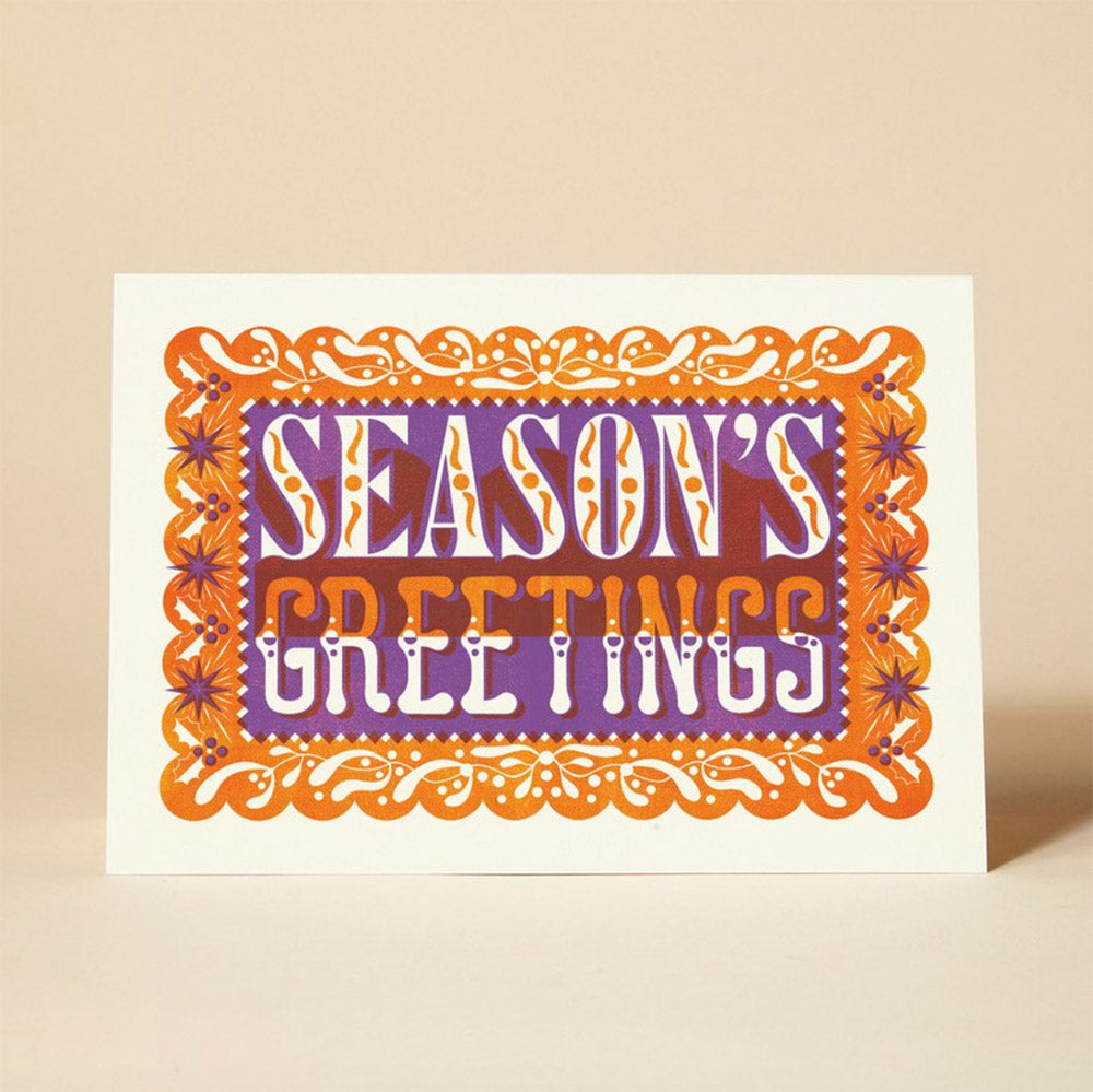 A festive Christmas card featuring the phrase "SEASON'S GREETINGS" in orange and purple lettering. The card has a decorative border with stars and snowflakes.