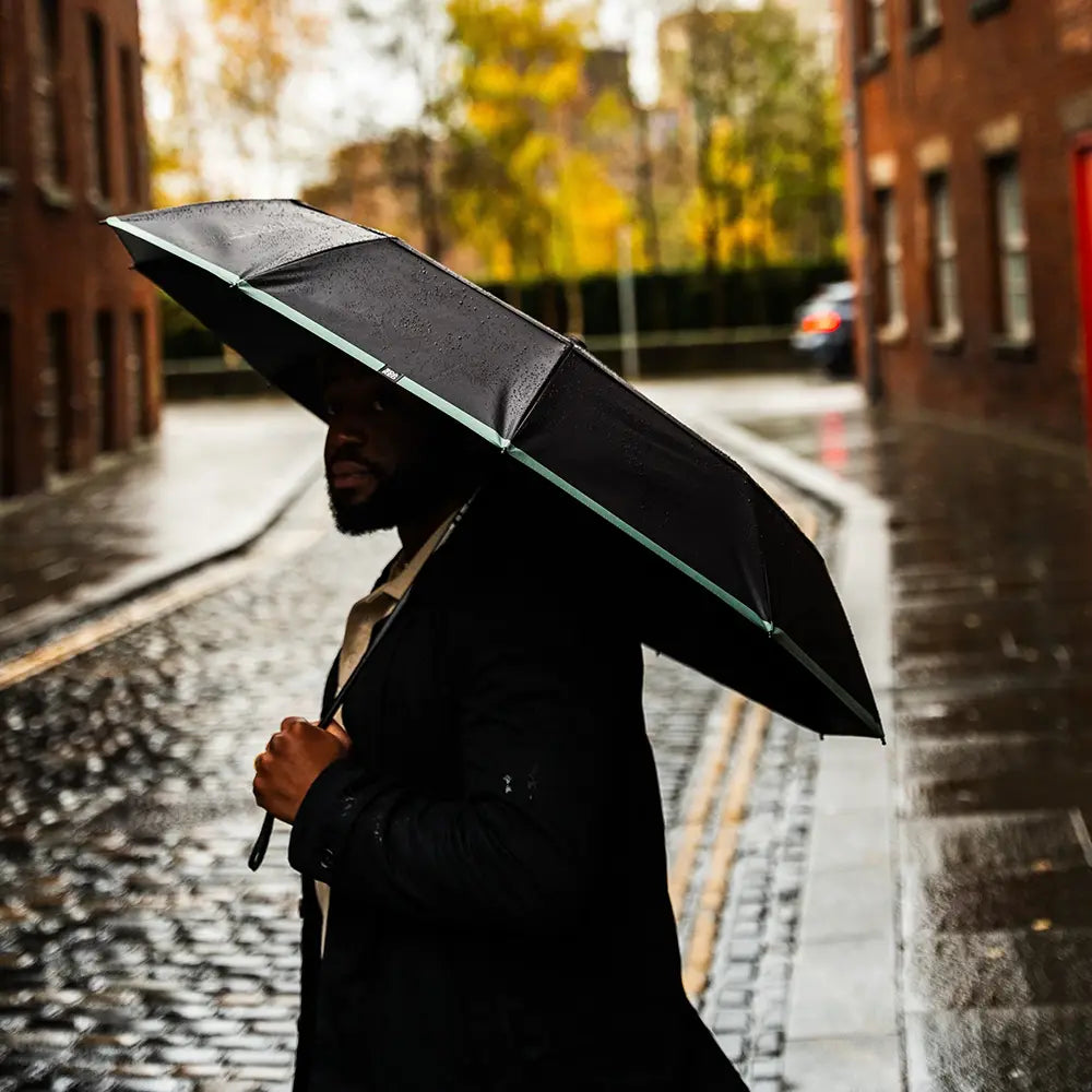 Person holding a black umbrella on a rainy street with brick buildings and a car in the background.