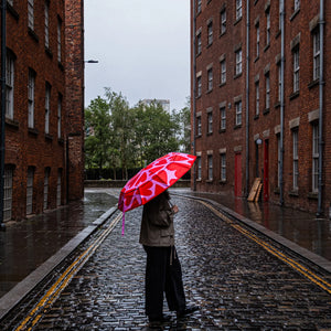 Person holding a red umbrella with a love heart pattern walking down a rain-soaked street between brick buildings.