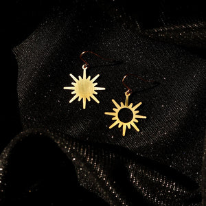 A pair of gold sun earrings with a minimalist design. The earrings feature a simple, circular outline with radiating sun rays. One is filled in and the other is hollow at the centre.