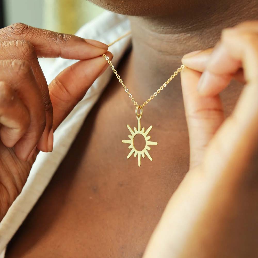 A close-up photo of a person wearing a gold sun pendant necklace. The person is using their fingers to hold the necklace.