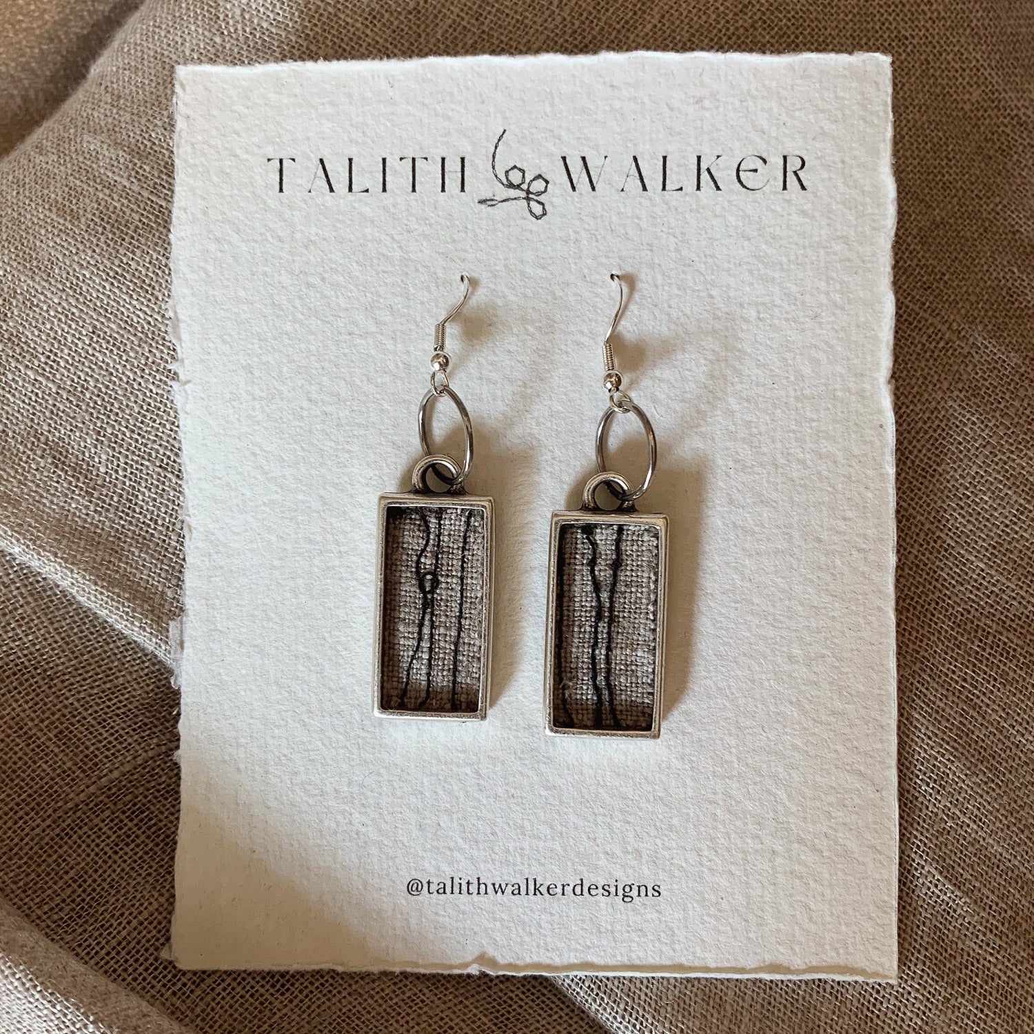 This image shows a pair of rectangular earrings with a textured, greyish fabric inside, hanging from silver hooks on a cream-coloured card. Placed on a cream fabric surface.