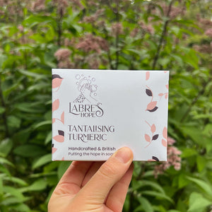 A person's hand the packaging for a bar of soap by Labre's Hope, against a garden backdrop.