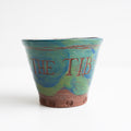 This is a close-up image of a small, decorative flower pot with the words "THE TIB" written on it, featuring a swirled green and blue design, against a plain background.
