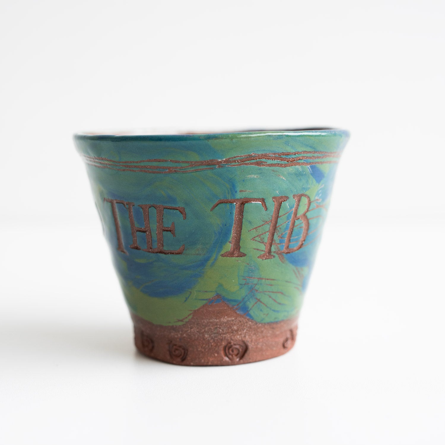 This is a close-up image of a small, decorative flower pot with the words "THE TIB" written on it, featuring a swirled green and blue design, against a plain background.