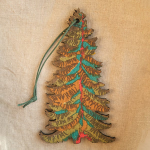 The image shows a wooden Christmas tree ornament. The ornament is decorated with intricate patterns in shades of green, orange, and red. It has a string attached to the top for hanging. 