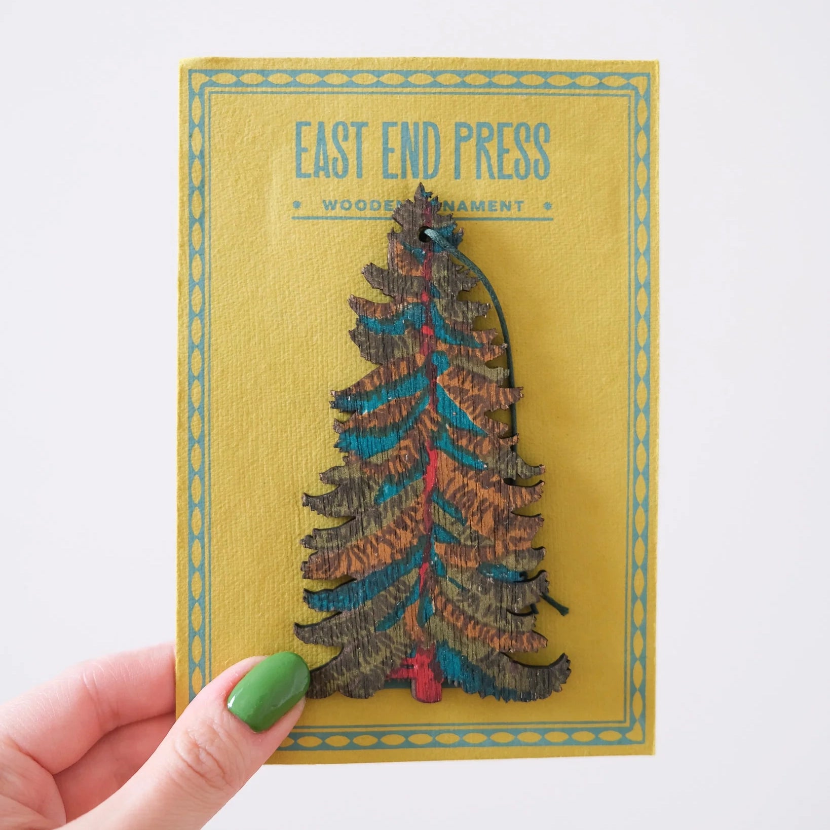 The image shows a person holding a wooden Christmas tree ornament in their hand. The ornament is decorated with colorful patterns and has a string attached to the top for hanging. It is packaged in a yellow card with the East End Press logo printed on it. 