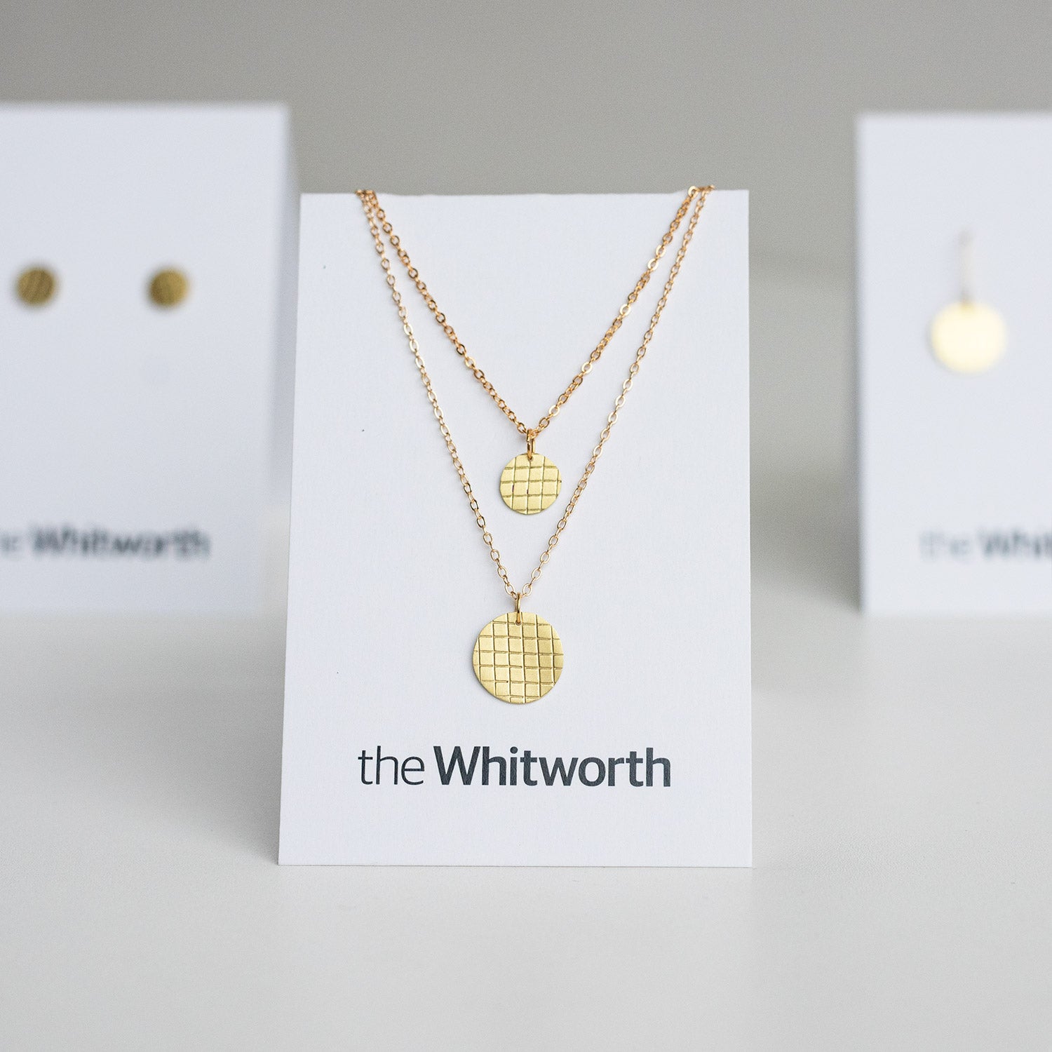 This image shows a gold necklace with a circular pendant displayed on a white card, with two other cards displaying gold earrings in the background.