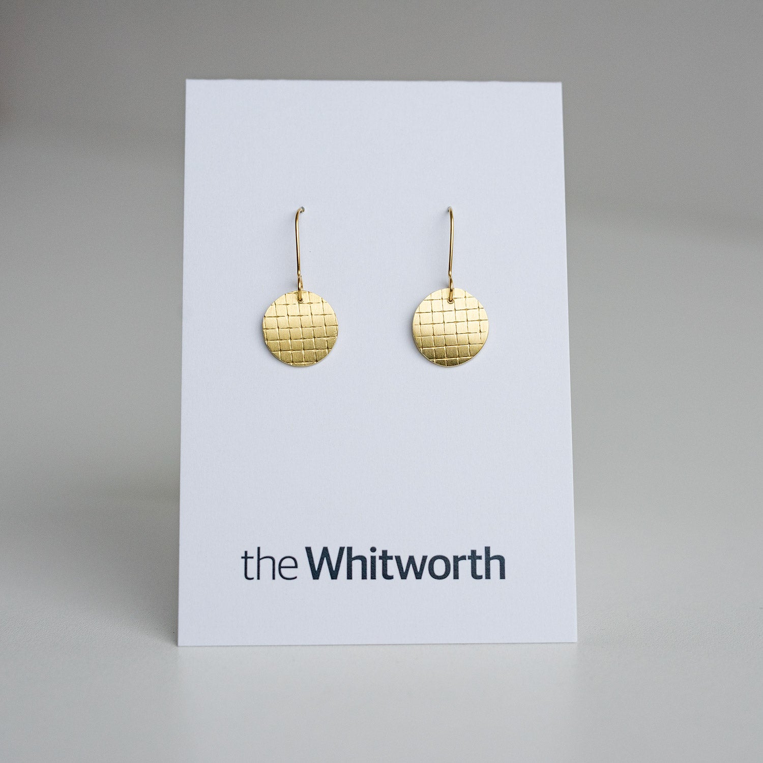 This image shows a pair of gold disc earrings with a grid pattern on a white card with "the Whitworth" printed on it.