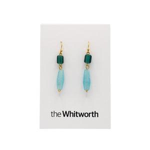 A pair of earrings with gold hooks, dark turquoise cubes, and faceted light blue teardrop beads hang on a white card with the Whitworth logo.