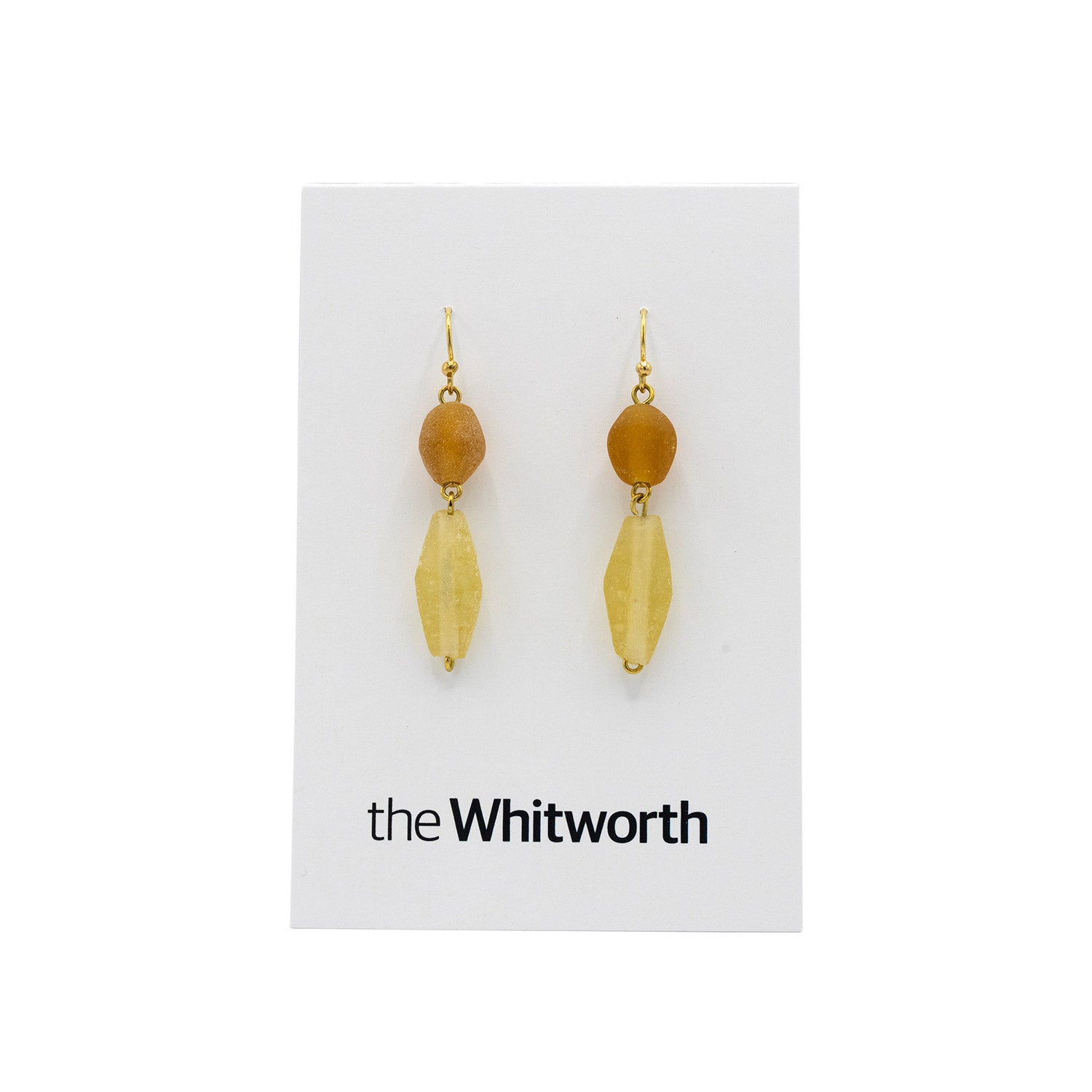 A pair of earrings with gold hooks, textured amber-coloured circles, and faceted teardrop-shaped amber beads hang on a white card with the Whitworth logo.