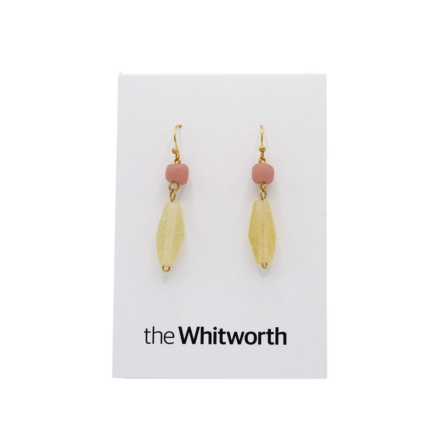 A pair of earrings with gold hooks, pink circles, and faceted teardrop-shaped light yellow beads hang on a white card with the Whitworth logo.