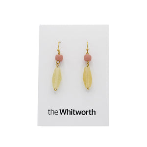 A pair of earrings with gold hooks, pink circles, and faceted teardrop-shaped light yellow beads hang on a white card with the Whitworth logo.