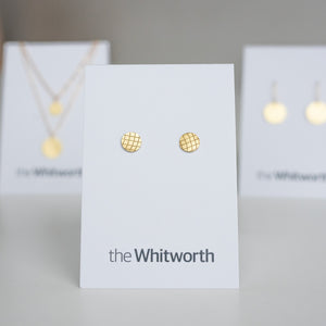 This image shows pair of stud earrings with grid-pattern etching displayed on a white card, with two other cards displaying gold jewellery in the background.