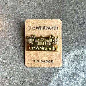 Gold bee pin badge in the shape of the Whitworth building, with the Whitworth logo. Against brown card backing on a marble surface.