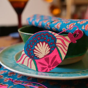 A photo of a beautifully decorated table setting. A hand-painted wooden ornament hangs from a napkin ring on a plate. The ornament features a colourful pattern of blue, pink, and green. There is a bowl and a glass on the table.