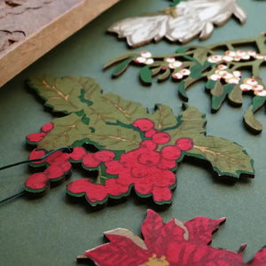 A close-up photo of hand-painted wooden Christmas ornaments. The ornaments depict various holiday plants, including holly with red berries, mistletoe with white berries, and a poinsettia. The ornaments are arranged on a green surface.