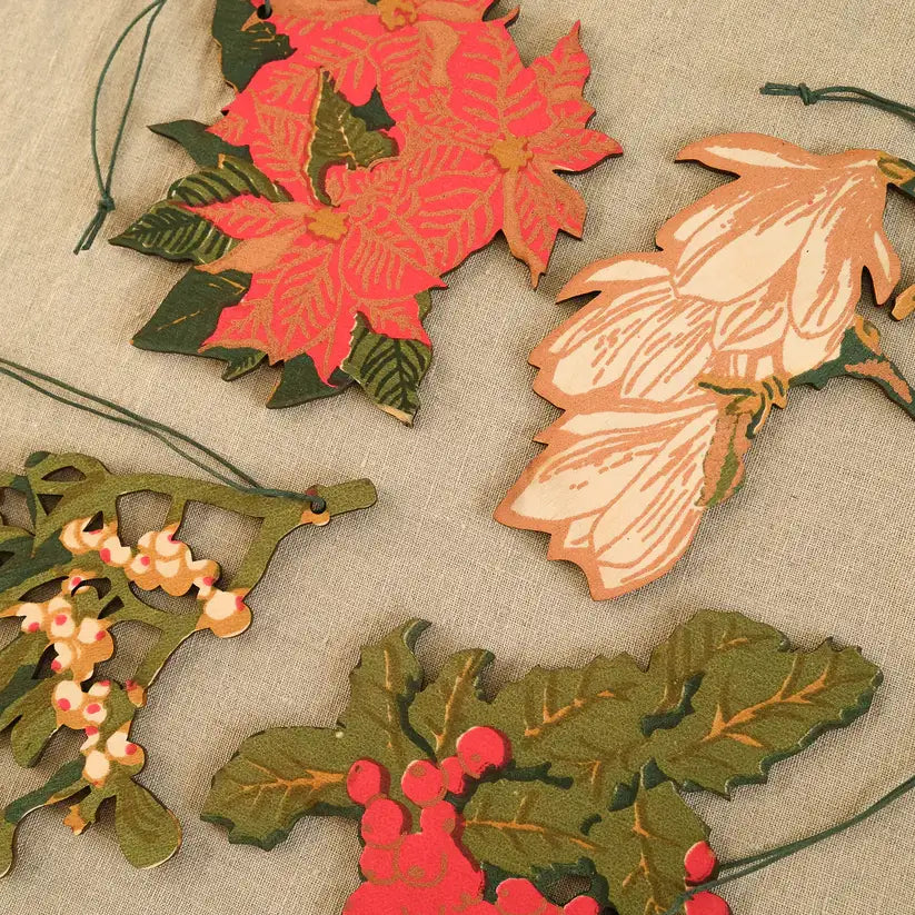 A photo of several hand-painted wooden Christmas ornaments arranged on a fabric surface. The ornaments depict different holiday plants, including poinsettias, magnolia leaves, and holly with berries.