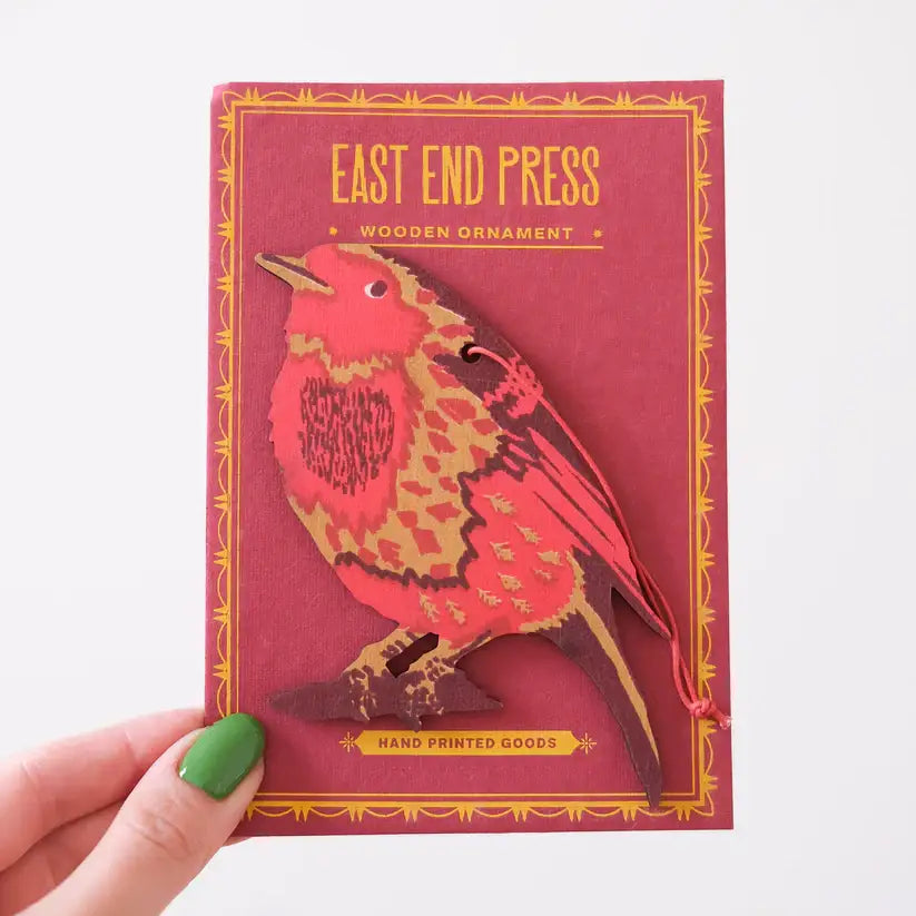 A photo of a person holding a red and gold packaging for a wooden Christmas ornament. The ornament is shaped like a robin bird and is hand-printed. The packaging features the brand and product name.