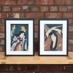 Two Japanese woodcut prints in black frame photographed in front of a brick wall