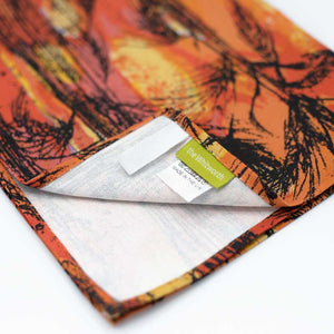 Detail of the tea towel with the golden harvest artwork lying on a white background. the tea towel is folded with one corner flipped up to show the white cotton rear side and Whitworth branding label.