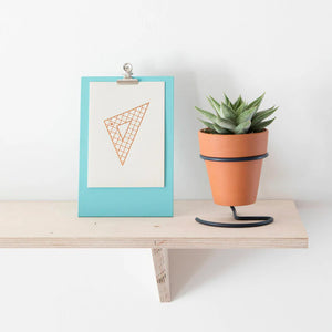 Lifestyle shot of the light blue clip board frame placed on a plywood shelf on a white wall. Beside the clipboard there is a terracotta planter with a succulent.