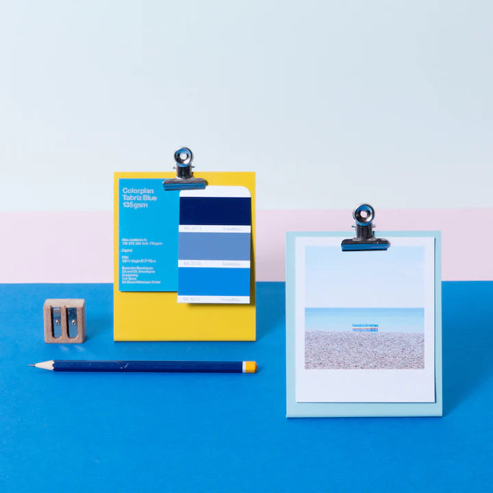Lifestyle shot on a bright blue surface with a light blue clipboard frame at the front and a yellow clipboard frame on the left and behind. A pencil and sharpener are respectively placed in front of beside the yellow frame.