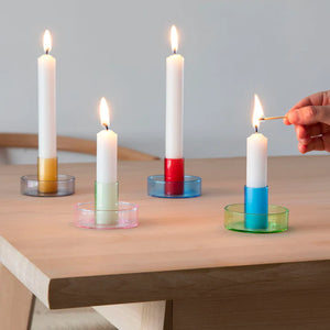 Lifestyle shot of four duo tone candle holders with a white, lit candle in each. A hand is reaching in from the right, holding a flaming match to the green and blue candle holders candle.