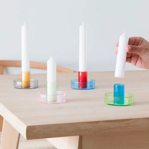 Lifestyle shot of four duo tone candle holders with a white, candle in three of them. The candles are on a wooden table and a hand is reaching in from the right, placing or removing a candle in the blue and green holder.