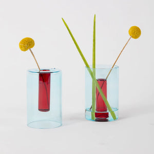 Two of the blue and red reversible vases shown in both possible options. Three grass stalks are inserted in the righthand vase while a single is in the lefthand one.