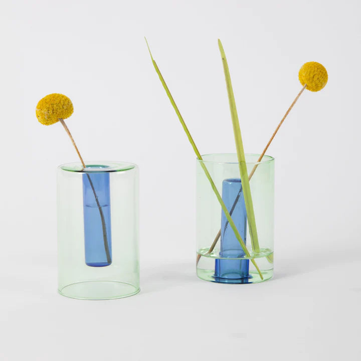Two of the blue and green reversible vases shown in both possible options. Three grass stalks are inserted in the righthand vase while a single is in the lefthand one.