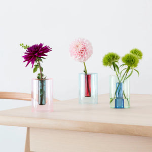Lifestyle shot of three reversible vases on a wooden table. The vase on the left has a dark purple flower while the one in the middle has a pale pink and the one on the right has three fluffy green seed pods. 