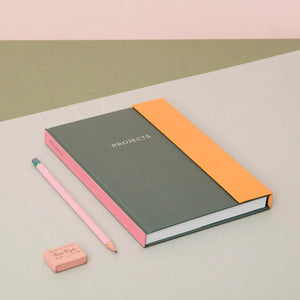 Lifestyle with the notebook placed on a grey surface with beige in the background. A pink pencil and a pink eraser are to the left of the notebook.