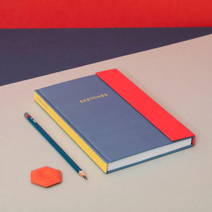 Lifestyle with the notebook placed on a grey surface with navy in the background. A teal pencil and a red eraser are to the left of the notebook.