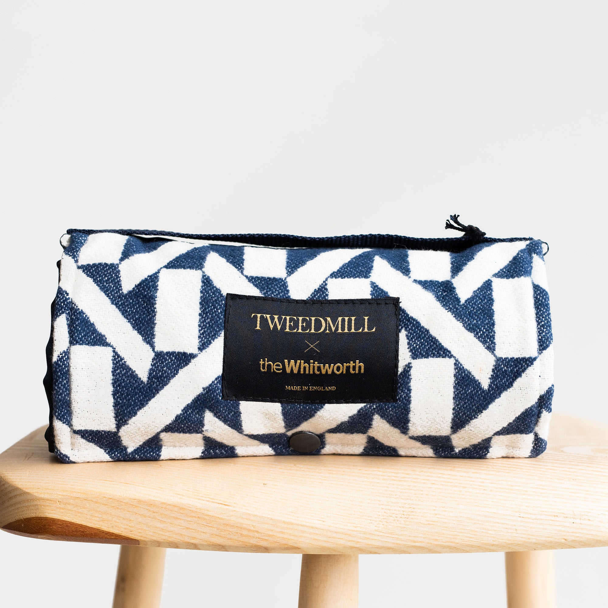 Tweedmill picnic rug rolled up and photographed on a stool. The rug has a navy and white abstract pattern design with the Tweedmill x Whitworth branding at the centre