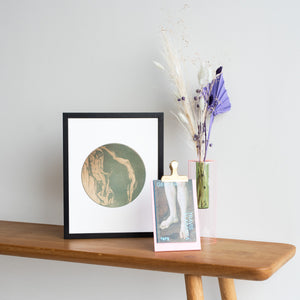 Lifestyle show of a print of The Coral Divers, a book about gender and a pink vase with some dried flowers. All resting upon a brown table with a white backdrop