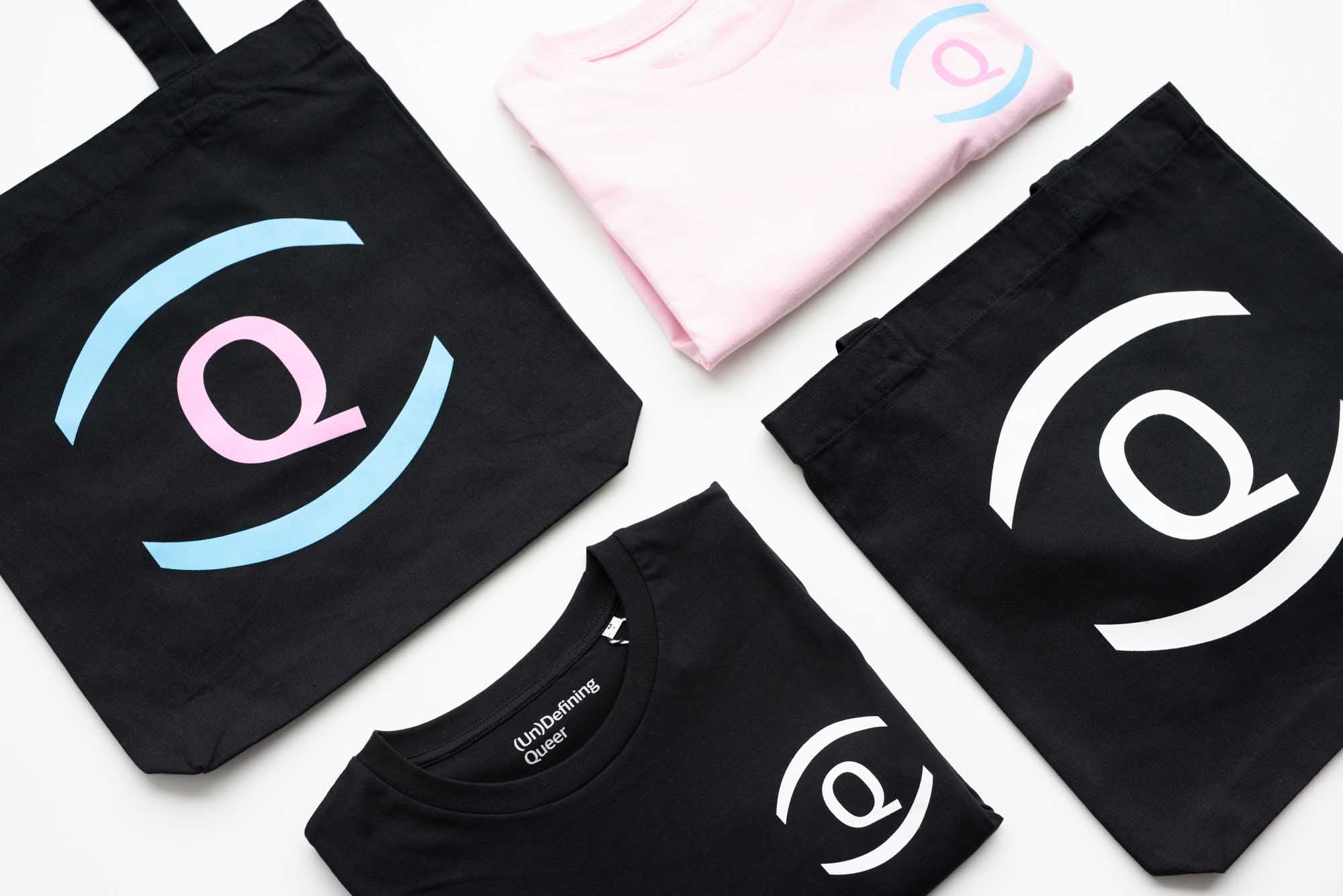 Flatlay image of pink t-shirts and tote bags folded on a white table with UnDefining Queer logo