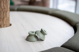 Lifestyle shot on a grey table. Pair of round, short candle holders in sage green. One holder is tilted on the side, leaning on the other.