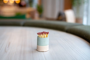 Lifestyle shot on a grey table. Sage and light grey coloured match pot with matches.