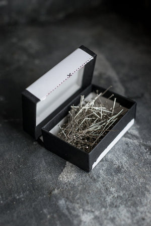The open card box of dress making pins on a dark slate surface.